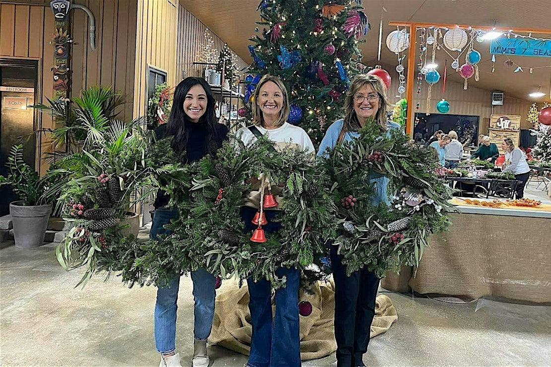Three women holding Christmas wreaths in a festive indoor setting with decorations and people in the background.